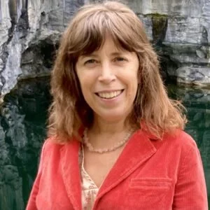 A woman with shoulder-length brown hair smiling, wearing a red blazer and a necklace, standing outdoors near rocky terrain.