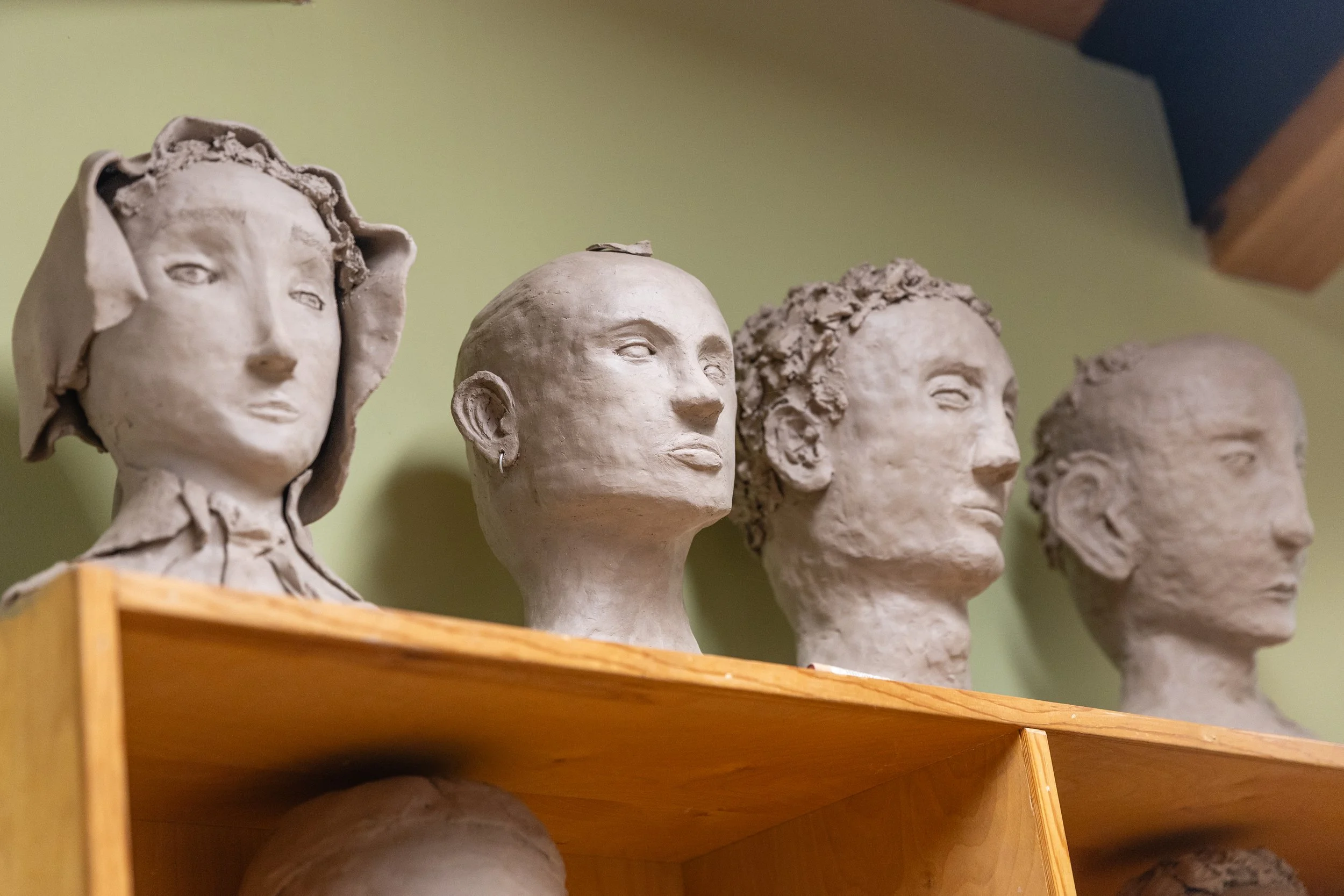 A wooden shelf holding multiple unfinished clay bust sculptures of human faces with various features and hairstyles.