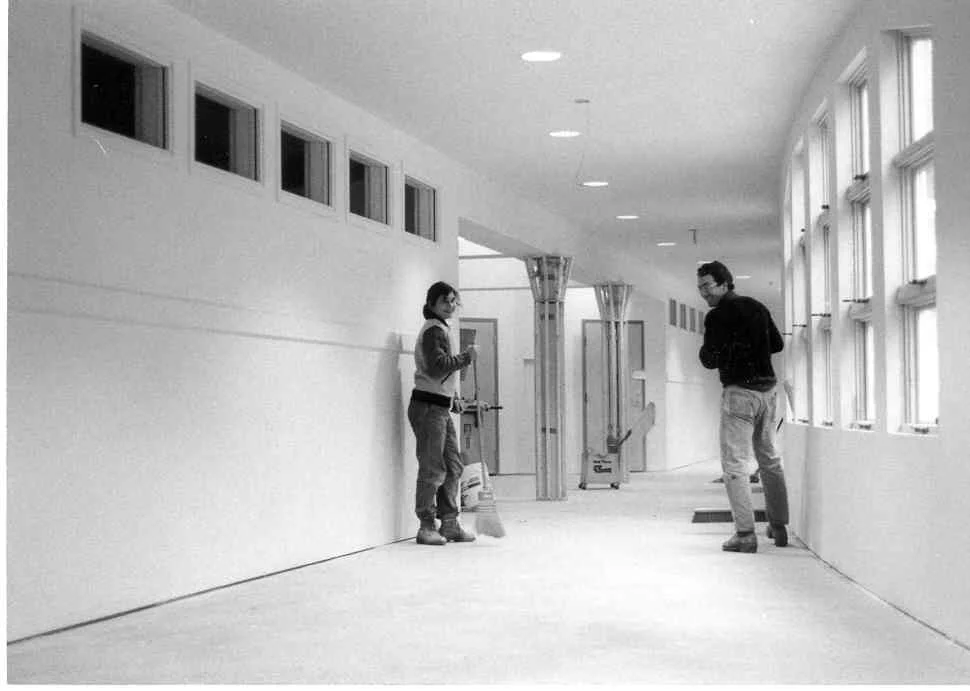 Two people, a woman and a man, are in a brightly lit corridor with large windows. The woman is wearing glasses and holding a cup, while the man is looking at her. There are cleaning supplies and some construction materials in the corridor, suggesting