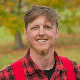 A man with light brown hair and a beard, smiling outdoors in a park with trees.
