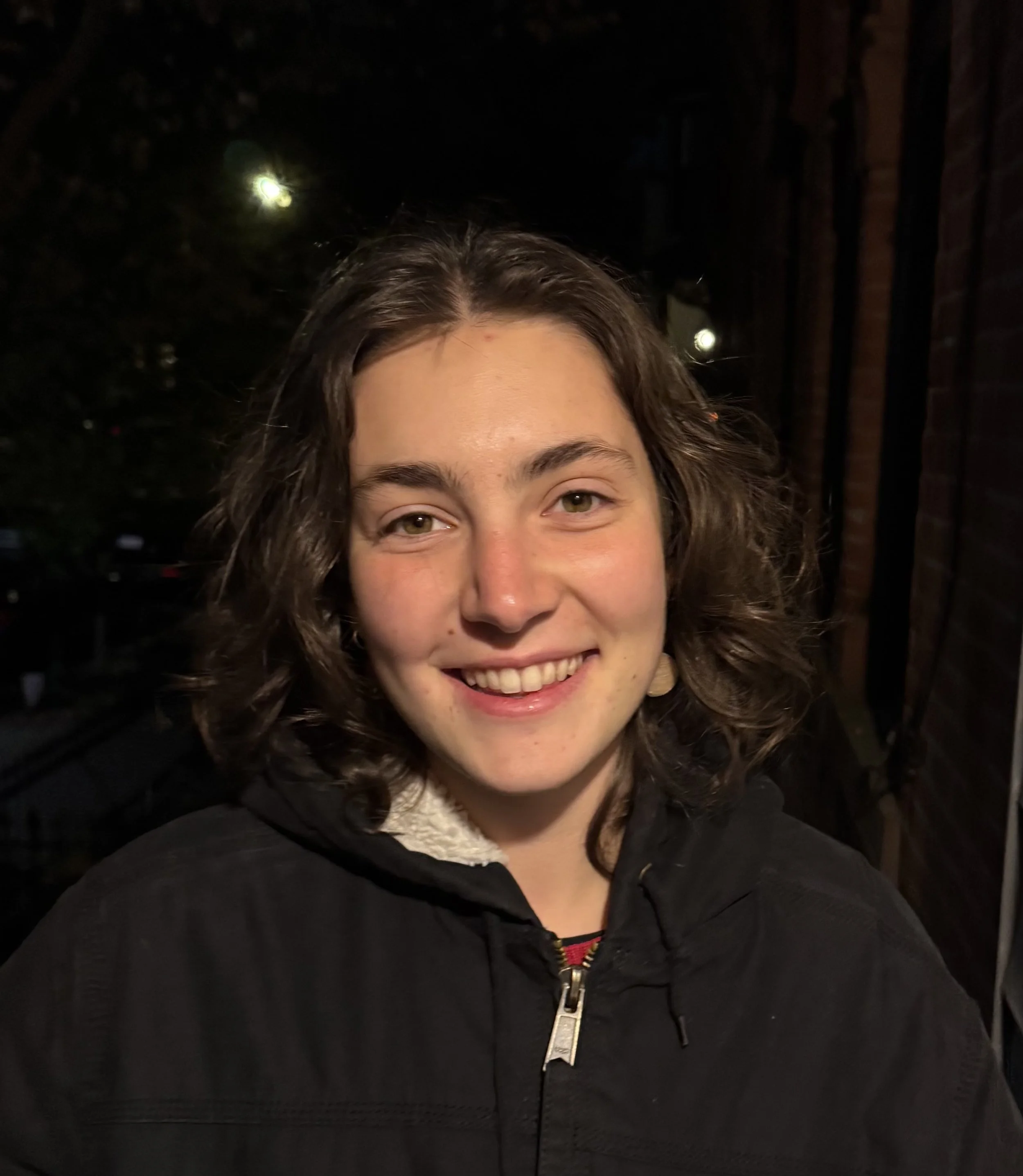 Close-up of a smiling young woman with curly brown hair, wearing a black jacket with a zipper, standing outdoors at night with dark trees and faint lights in the background.