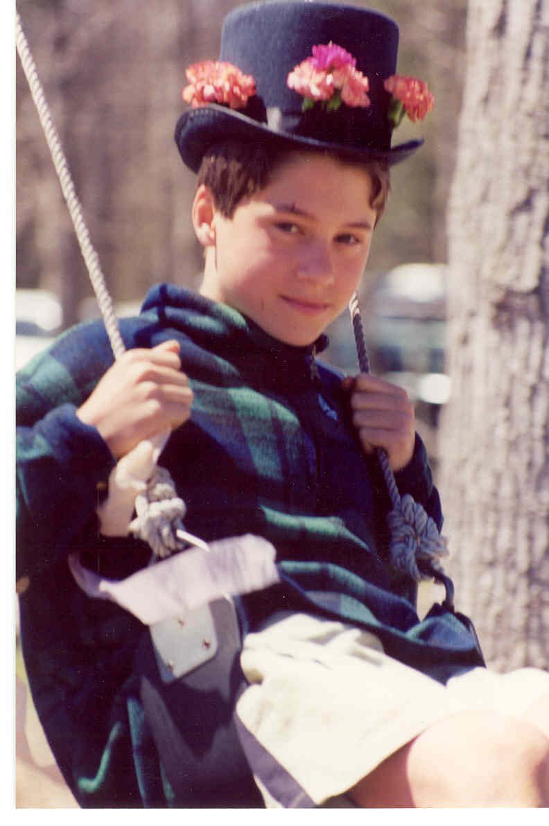A teenage student outdoors, wearing a black top hat decorated with pink and red flowers, sitting on a swing. He has a smile, and there are trees in the background.