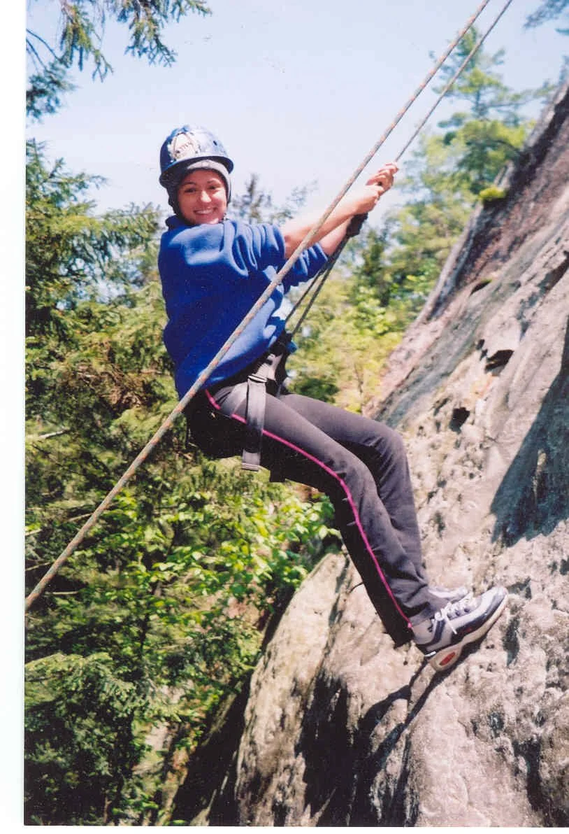 A young woman rock climbing outdoors, wearing a helmet, blue jacket, black pants with pink stripes, sneakers, and safety harness, holding onto the rock face with a rope.