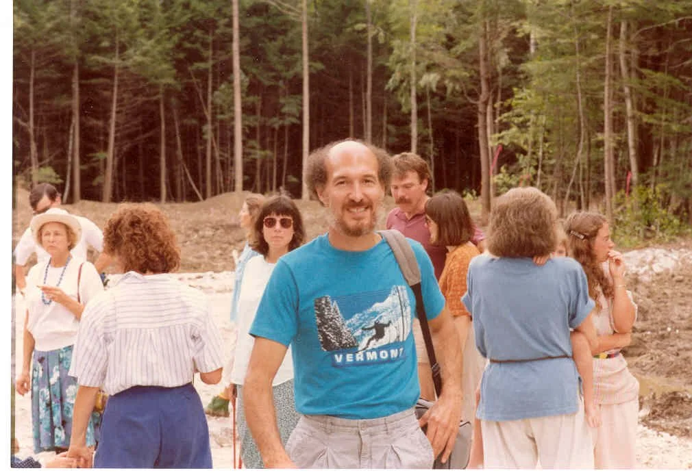 A group of people outdoors in a wooded area, some wearing summer clothes and hats, with a man in a blue shirt smiling at the camera in the foreground.