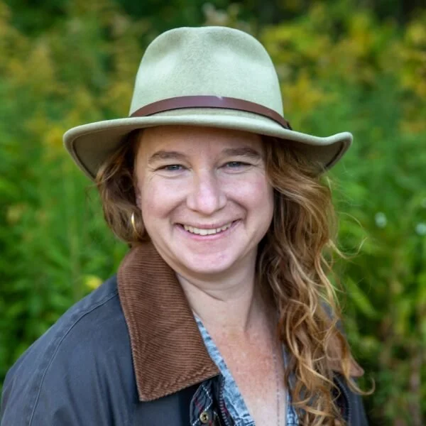 A woman with wavy red hair wearing a beige fedora hat with a brown band, a dark jacket, and a light blue shirt, smiling outdoors with green foliage in the background.