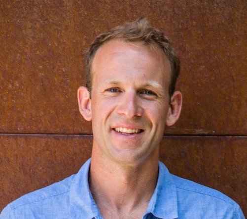 A smiling man in a blue collared shirt standing in front of a brown wooden wall.