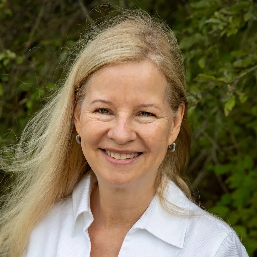 Smiling woman with long blonde hair wearing a white collared shirt and earrings outdoors with green foliage background.