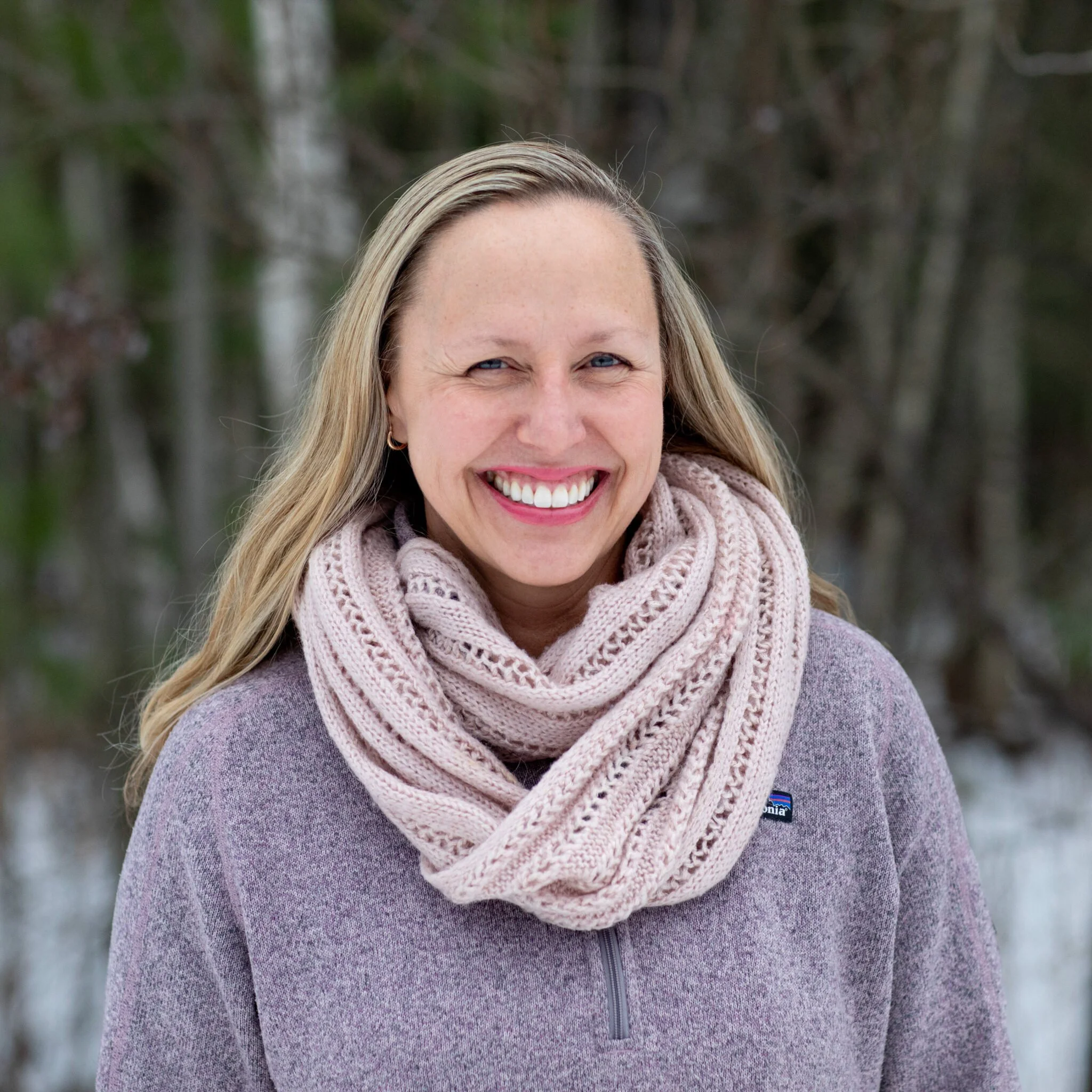 Close-up of a smiling woman with blonde hair wearing a pink knitted scarf and a purple sweater outdoors.