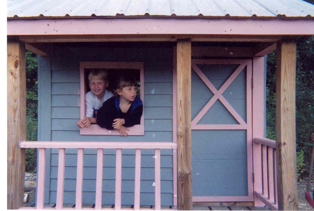 Two children looking out of a playhouse window, smiling and leaning on the windowsill.