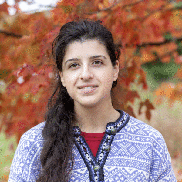 A young woman with long, dark hair and a light smile, wearing a patterned sweater, stands outdoors in front of red and orange autumn leaves.