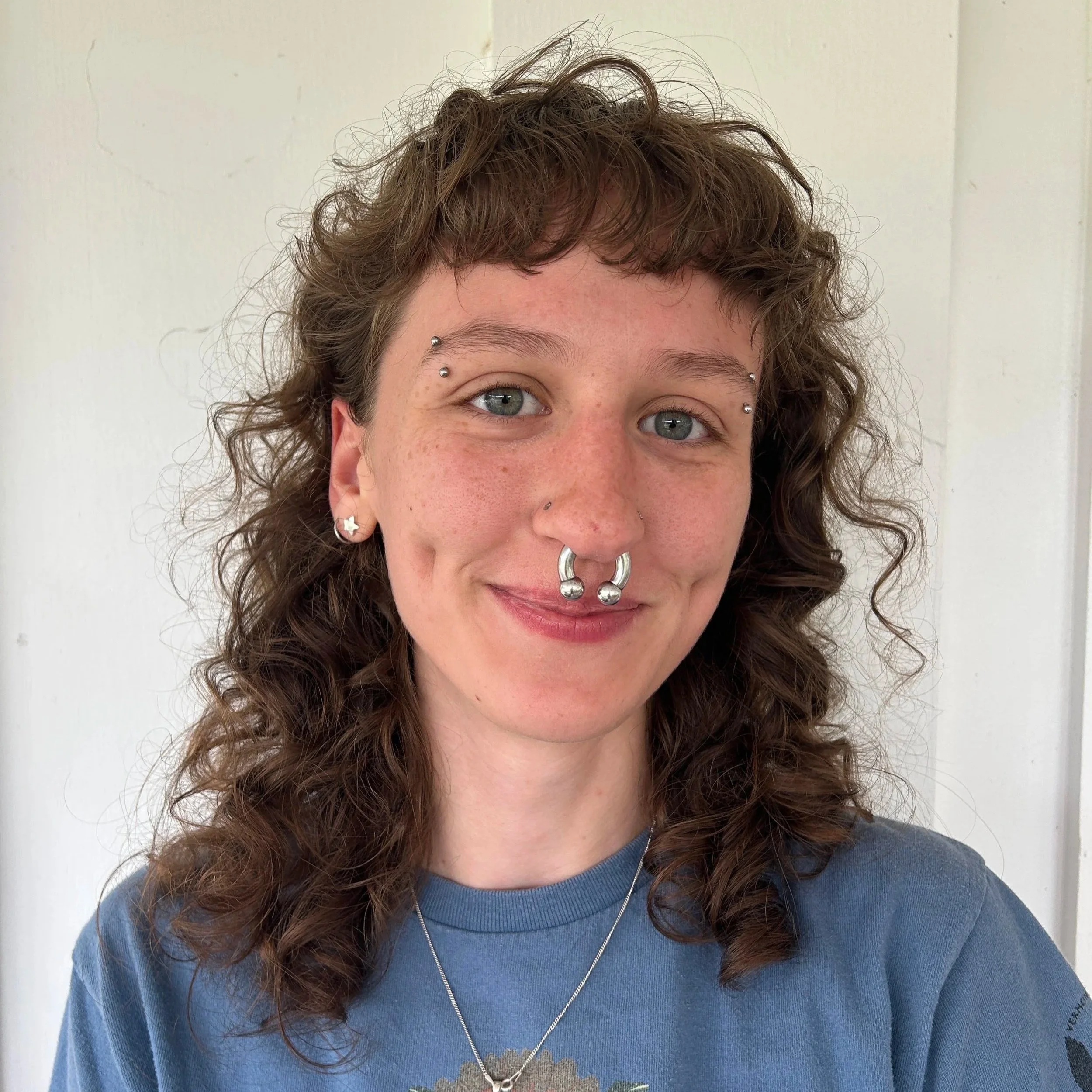 Part of a woman’s face showing her green eyes, nose, and curly brown hair, with facial piercings including nose, eyebrow, and ear jewelry, wearing a blue shirt and a necklace, standing against a plain white background.