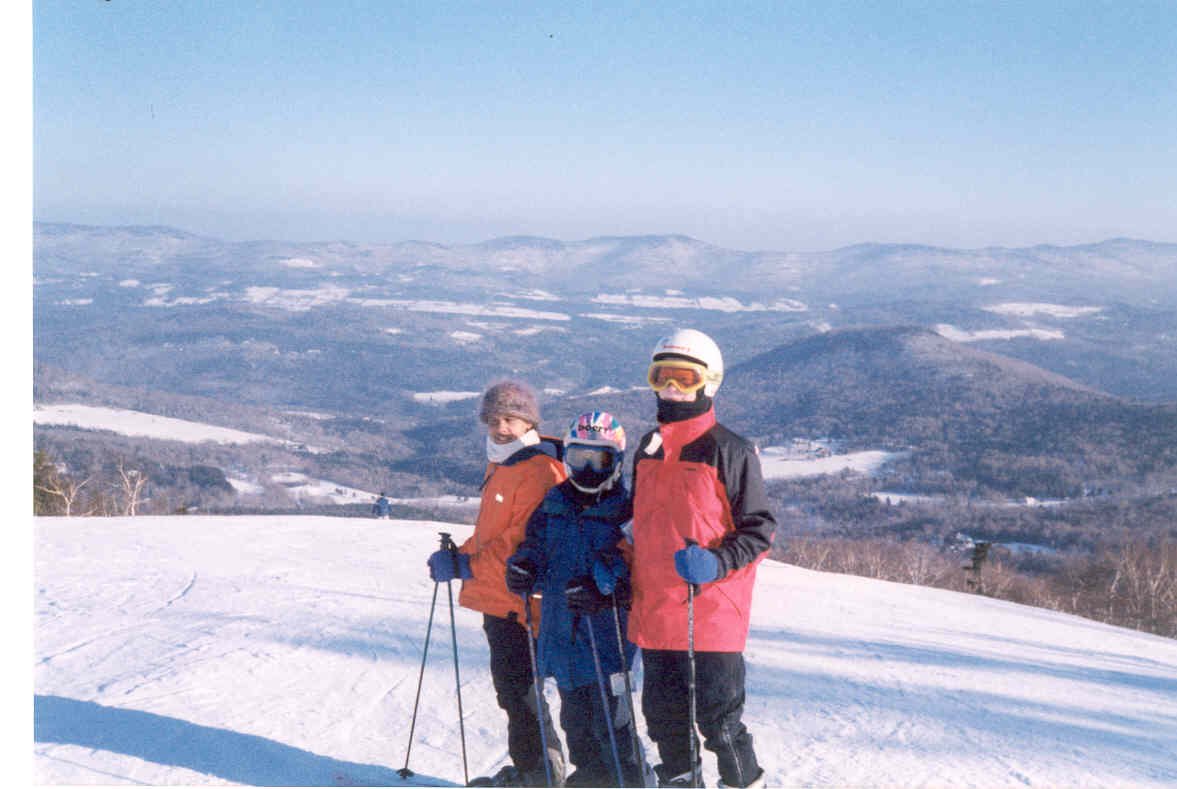 Three people in winter clothing with ski gear standing on a snowy mountain slope with a scenic view of snow-covered hills and valleys in the background.