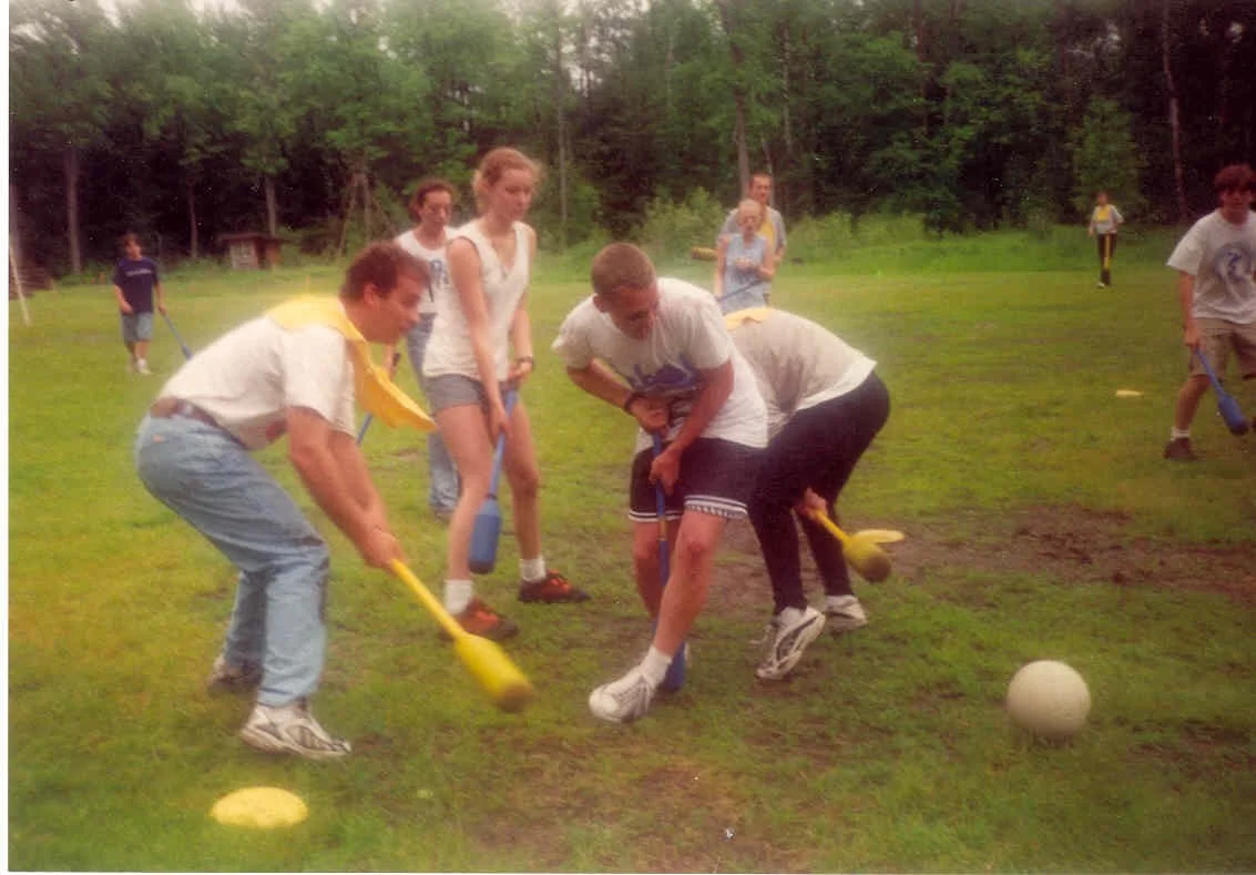 Group of young people playing a game with plastic shovels and a white ball outdoors on a grassy field, surrounded by trees.