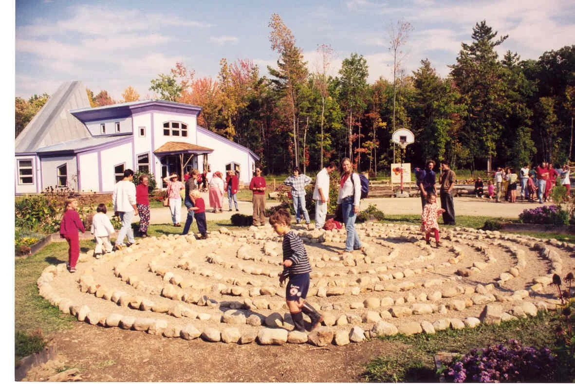 Children and adults playing on a stone labyrinth outdoors with a purple building in the background and trees with autumn foliage.