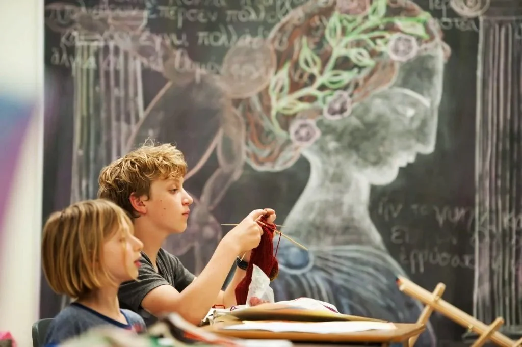 Boy knitting in front of a chalkboard with colorful art