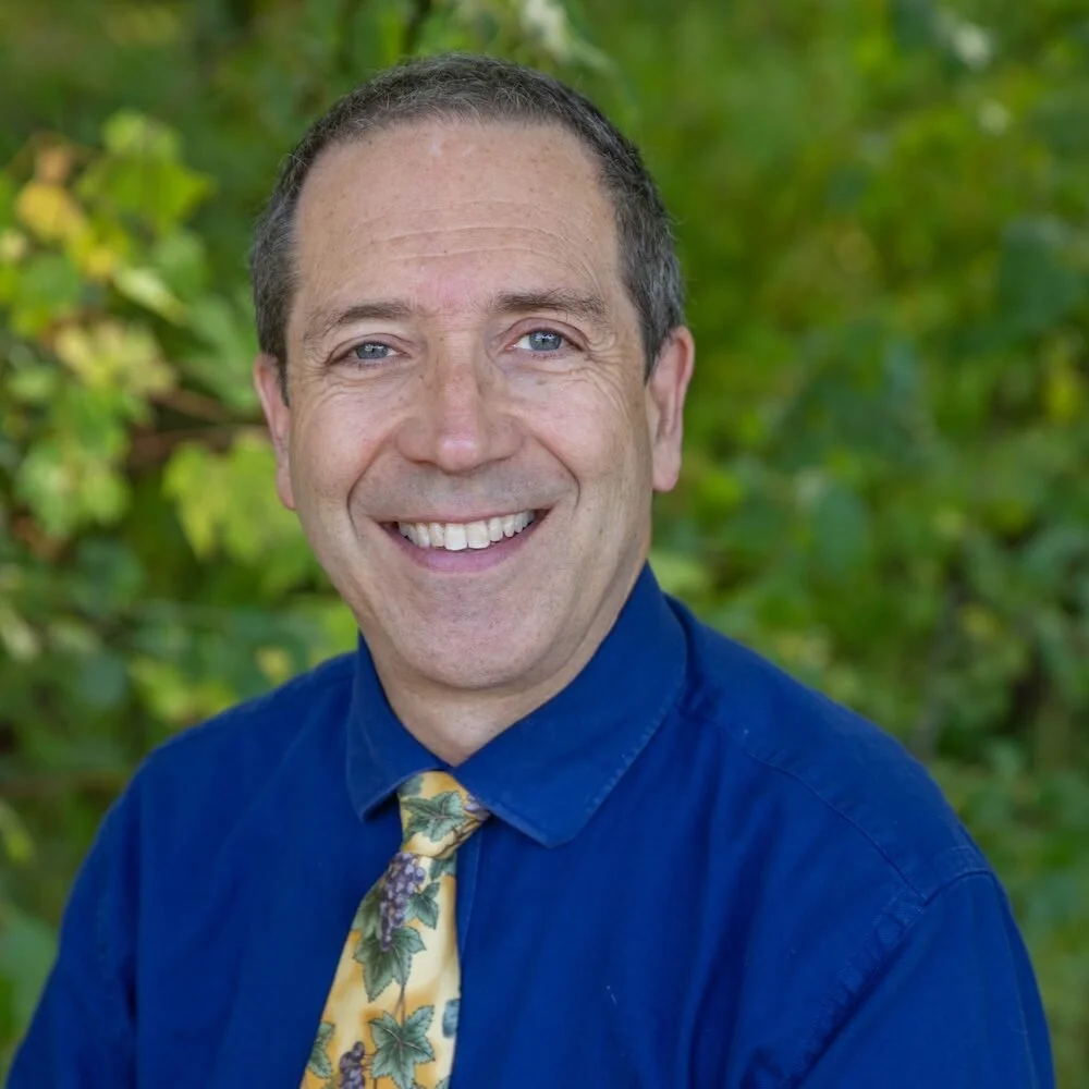 A smiling middle-aged man with short dark hair and blue eyes, wearing a blue dress shirt and a yellow tie with a grapevine pattern, standing outdoors with green foliage in the background.