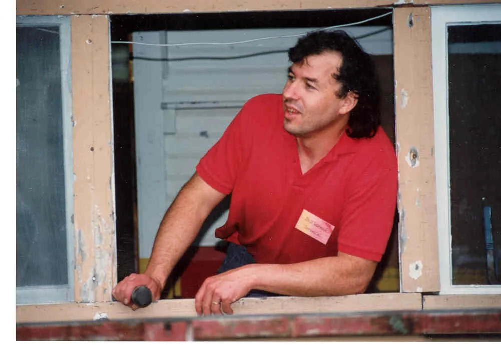 A man with curly dark hair, wearing a red shirt with a name tag, leaning out of a wooden window frame with chipping paint and looking to his left.