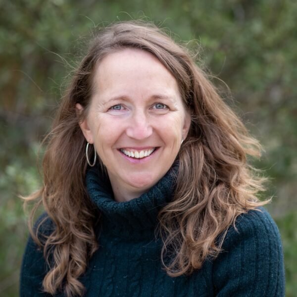 A woman with long, curly brown hair, blue eyes, and light skin, smiling outdoors with blurred greenery in the background, wearing a dark sweater and hoop earrings.