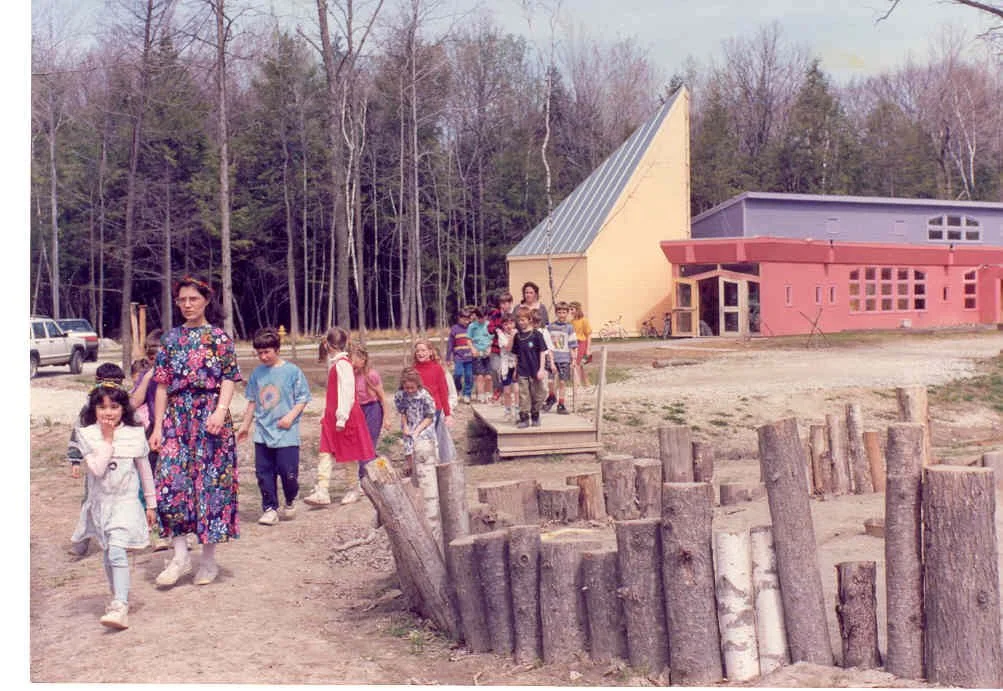Children and adults walking outdoors near a colorful building, trees in the background, and a wooden border along a dirt path.