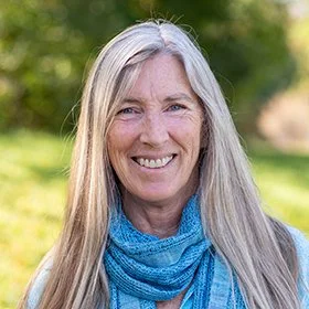 A woman with long gray hair smiling outdoors in a green park, wearing a blue scarf.
