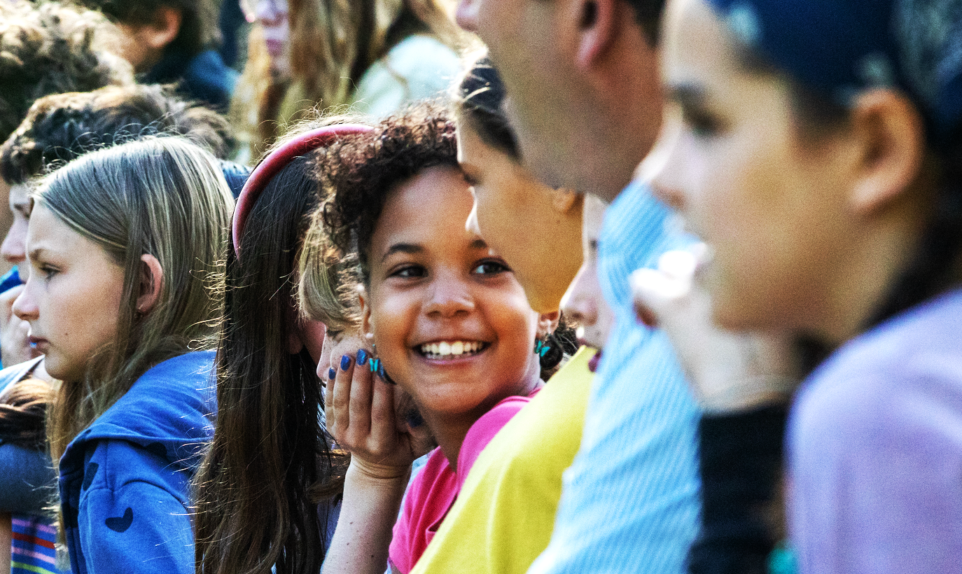 A group of diverse children sitting outdoors, with a girl smiling joyfully at the camera.