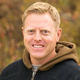 Close-up of a smiling man with short blond hair, wearing a black and brown jacket, outdoors with blurred autumn foliage in the background.