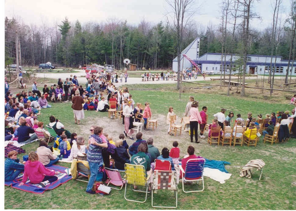 A large group of people gathered outdoors in a grassy area, watching a performance or event, with some sitting on chairs or blankets and others standing. There are children and adults, and a building and trees are visible in the background.