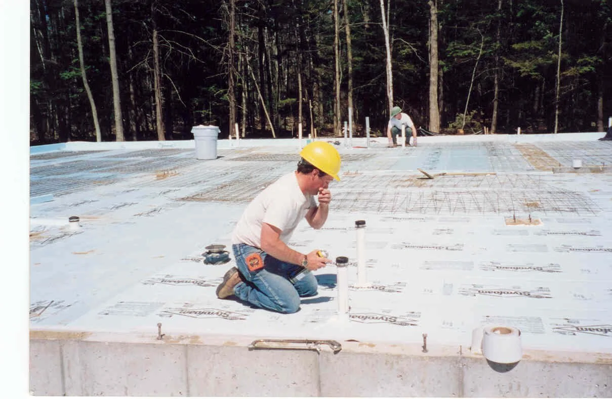 Construction workers installing insulation on a building foundation in a wooded area.