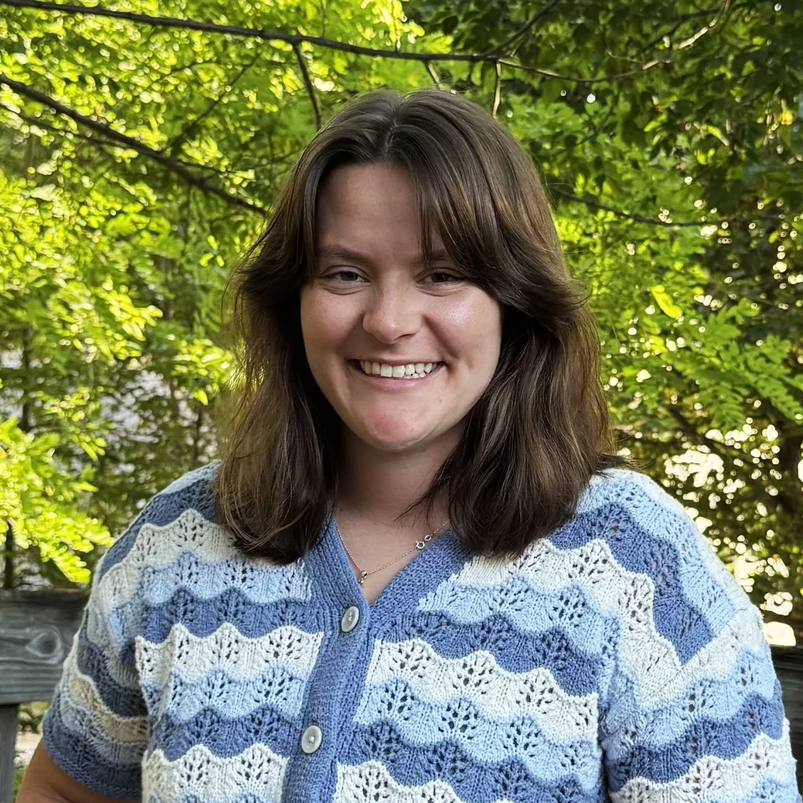A smiling woman with shoulder-length brown hair wearing a blue and white crocheted cardigan outdoors surrounded by green foliage.