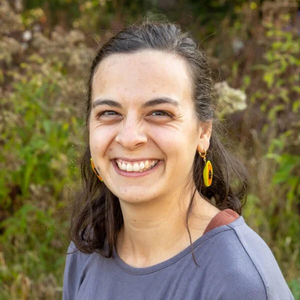 A young woman smiling outdoors with brown hair, wearing yellow earrings and a gray shirt, in front of green foliage.