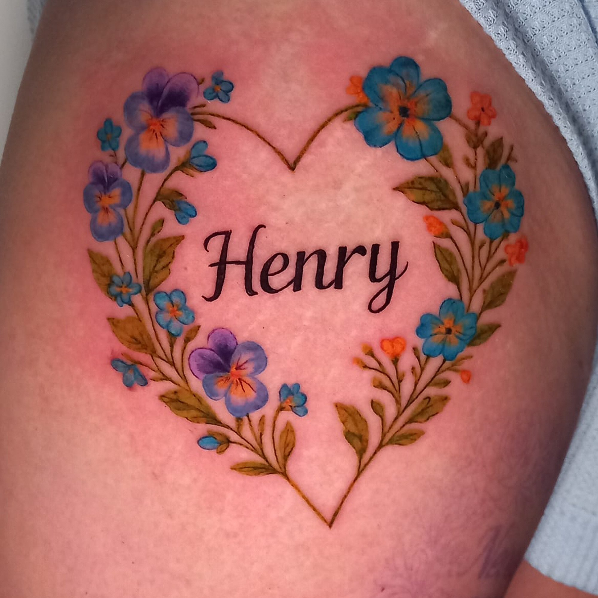 Close-up of a person's chest with a floral tattoo featuring blue and orange flowers arranged in a heart shape, with the word 'Honey' in the center.
