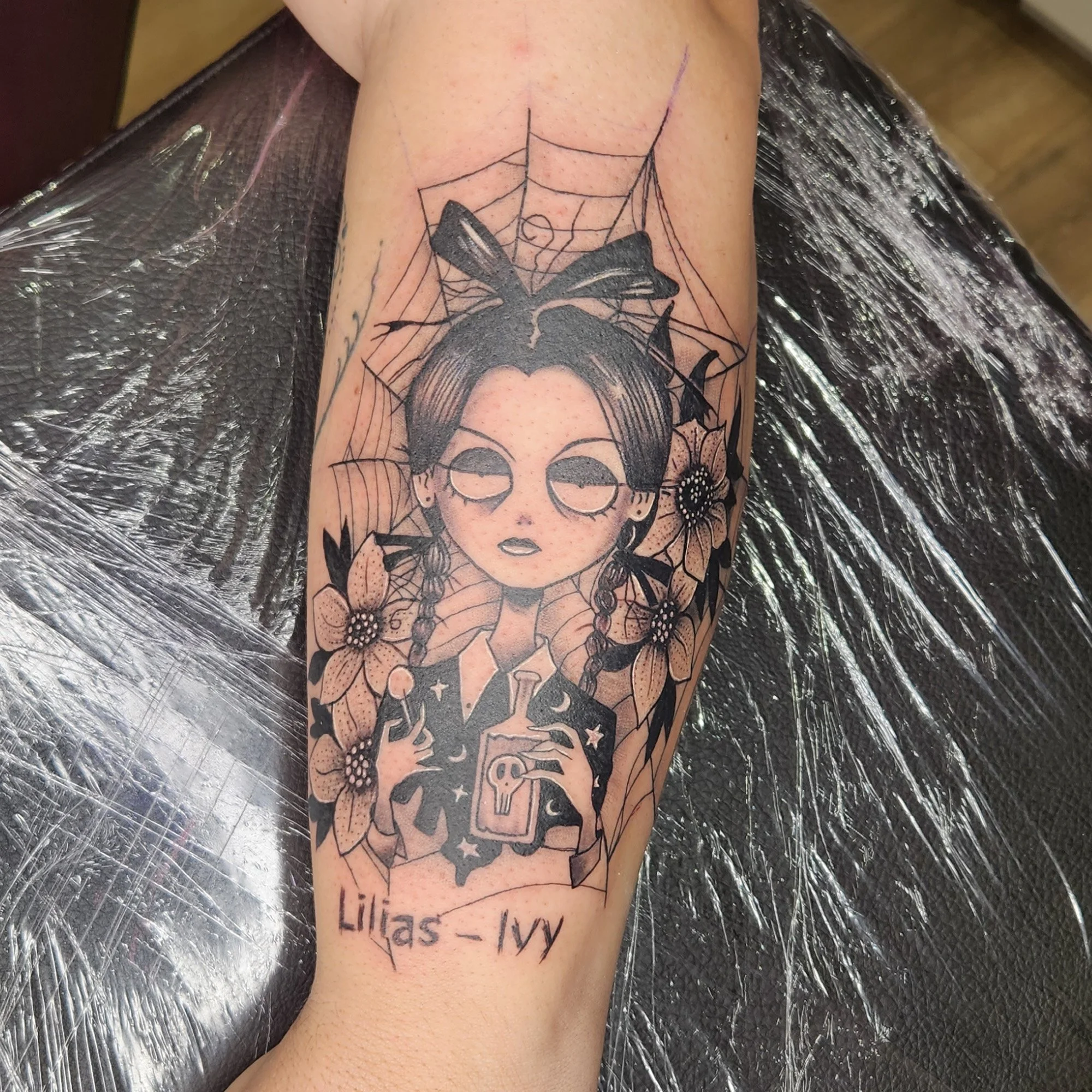 Tattoo on a person's arm of a cartoonish woman with glasses, short hair, and flowers, framed by a spider web design.