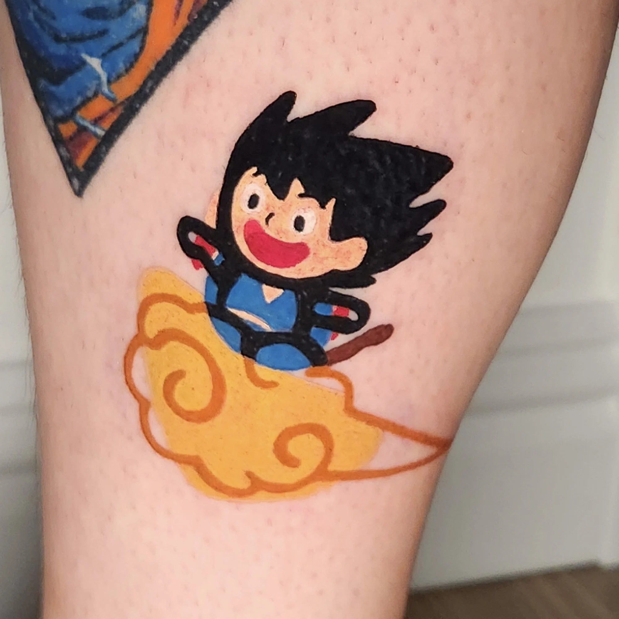Close-up of a colorful tattoo on skin featuring a cartoon character with spiky black hair, red mouth, and yellow teeth, surrounded by orange and yellow circular patterns.