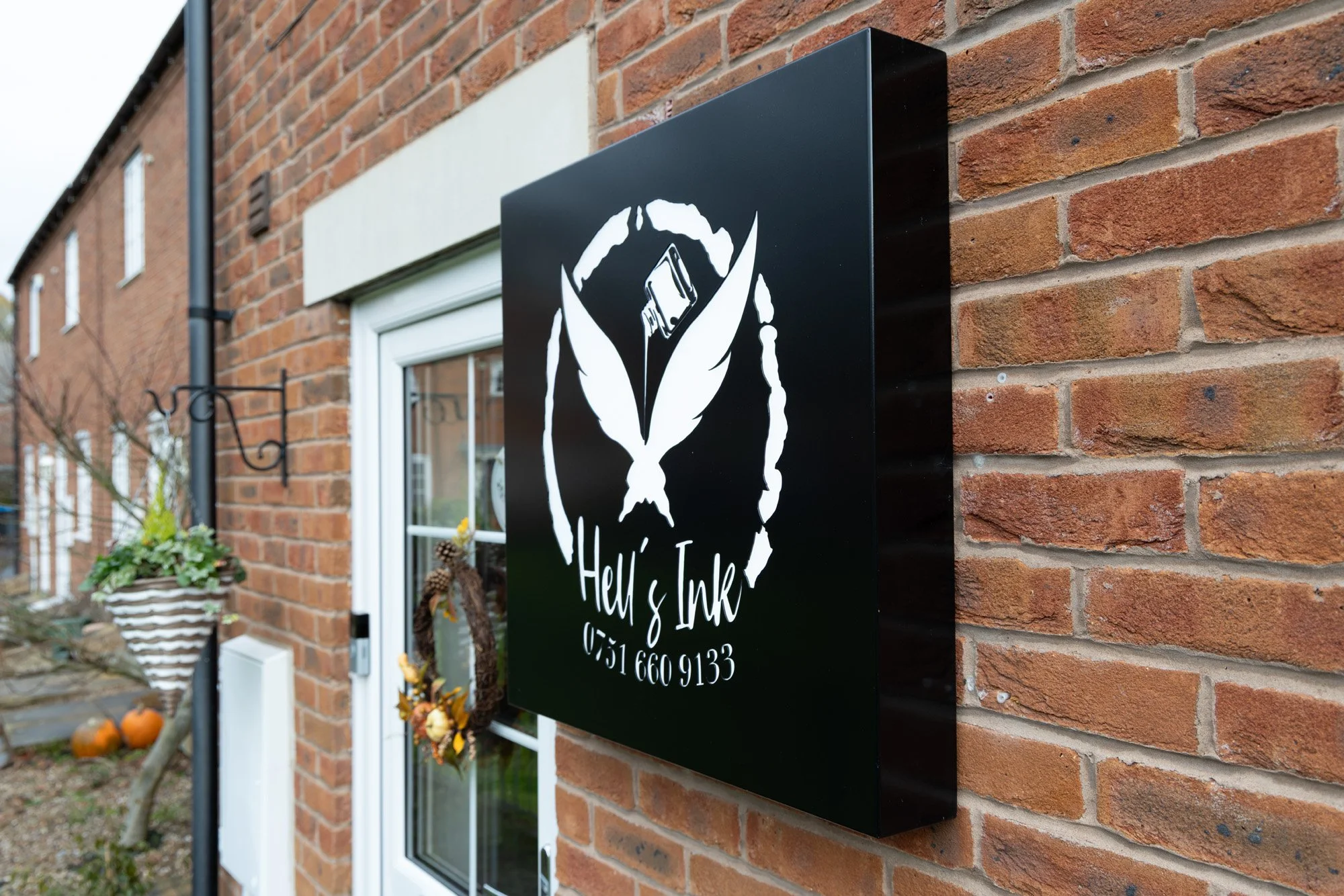 Black sign on a brick wall with white text and a distinctive logo for 'Helli & Ink', including a telephone number.