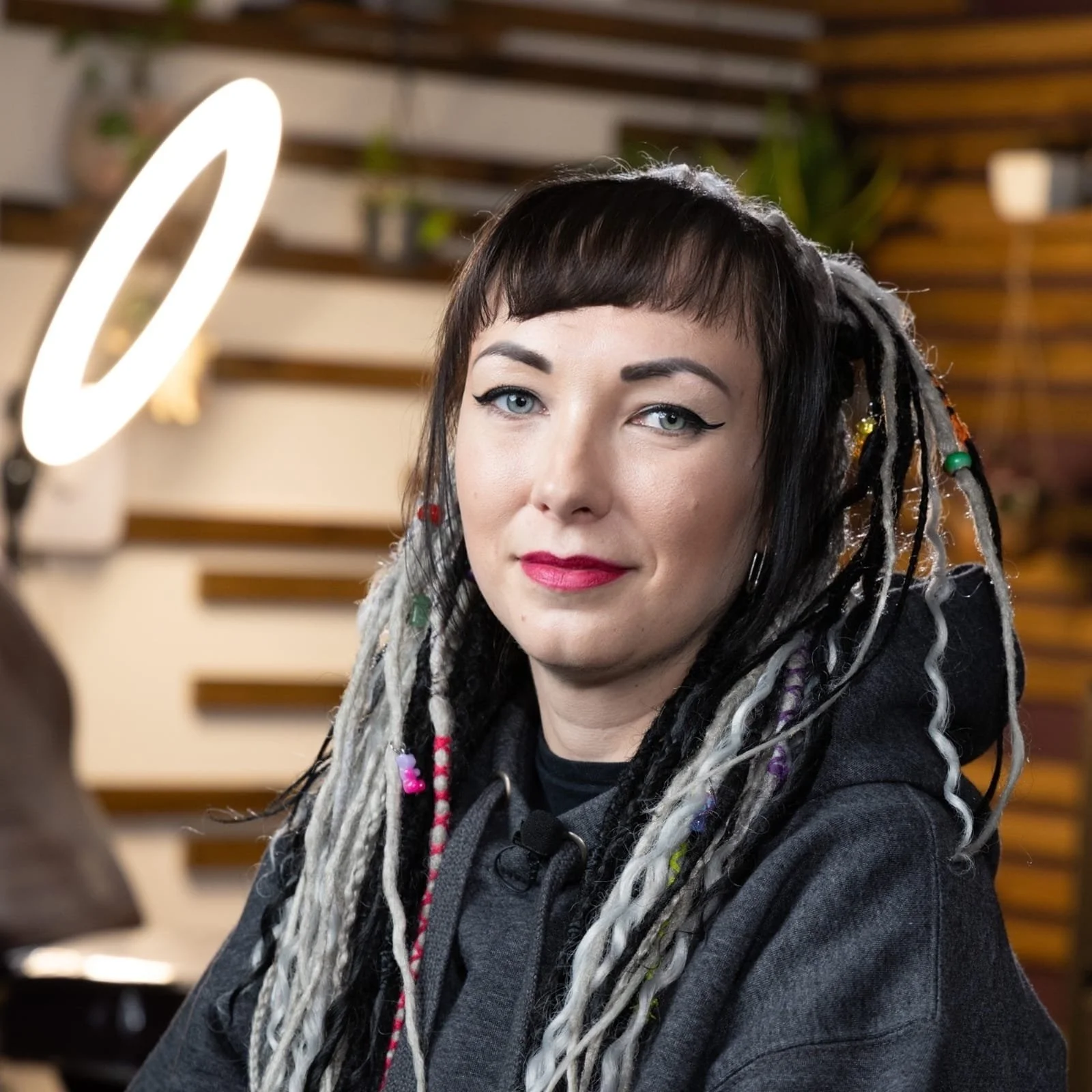 A woman with short dark hair, light makeup with winged eyeliner and red lipstick, wearing a gray hoodie, is sitting indoors in front of a wooden wall with greenery. There is a ring light to her left.