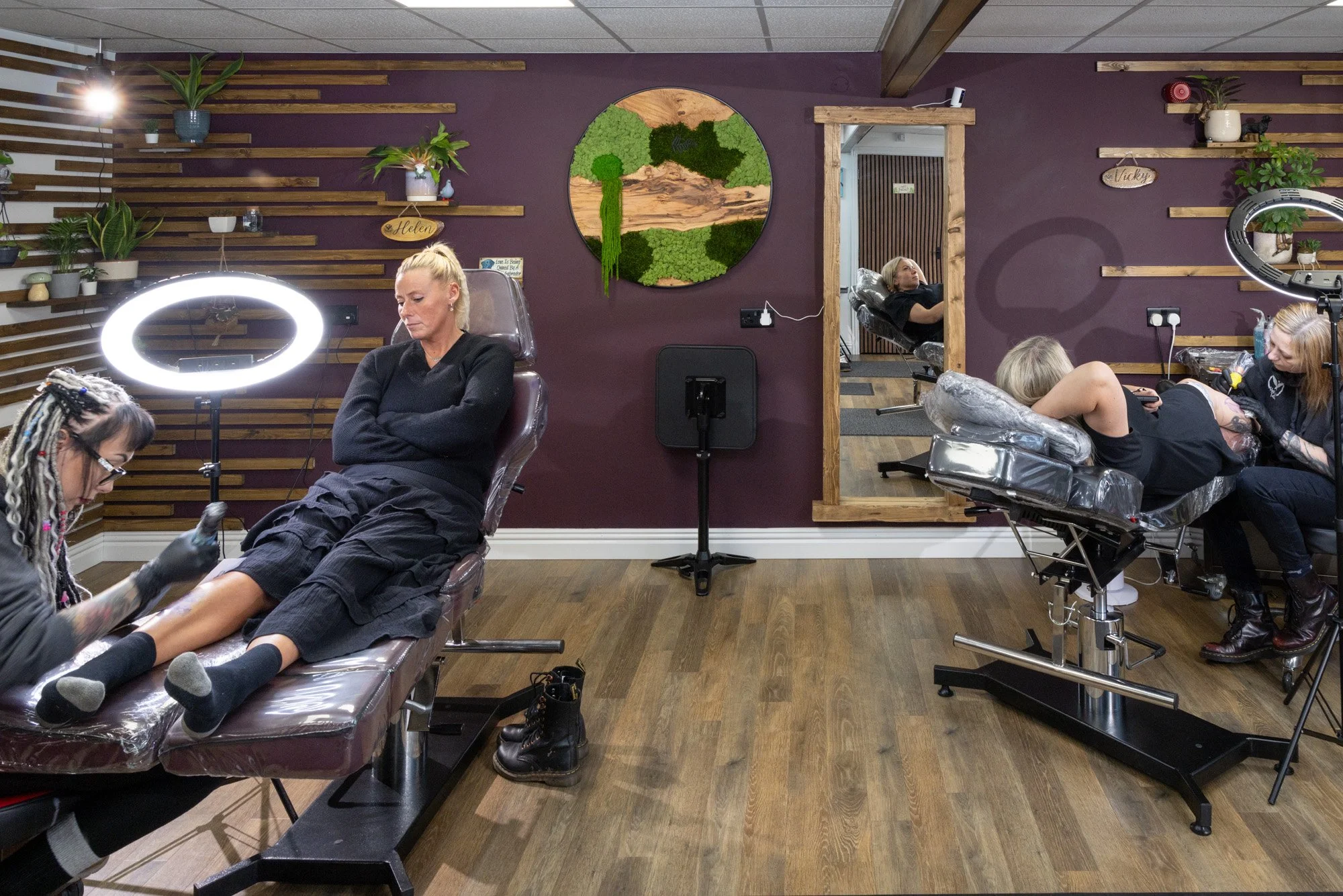 A woman receiving a pedicure at a nail salon, sitting in a reclined chair with a technician applying nail polish, and another woman relaxing in the background, all in a room decorated with plants and wooden wall accents.