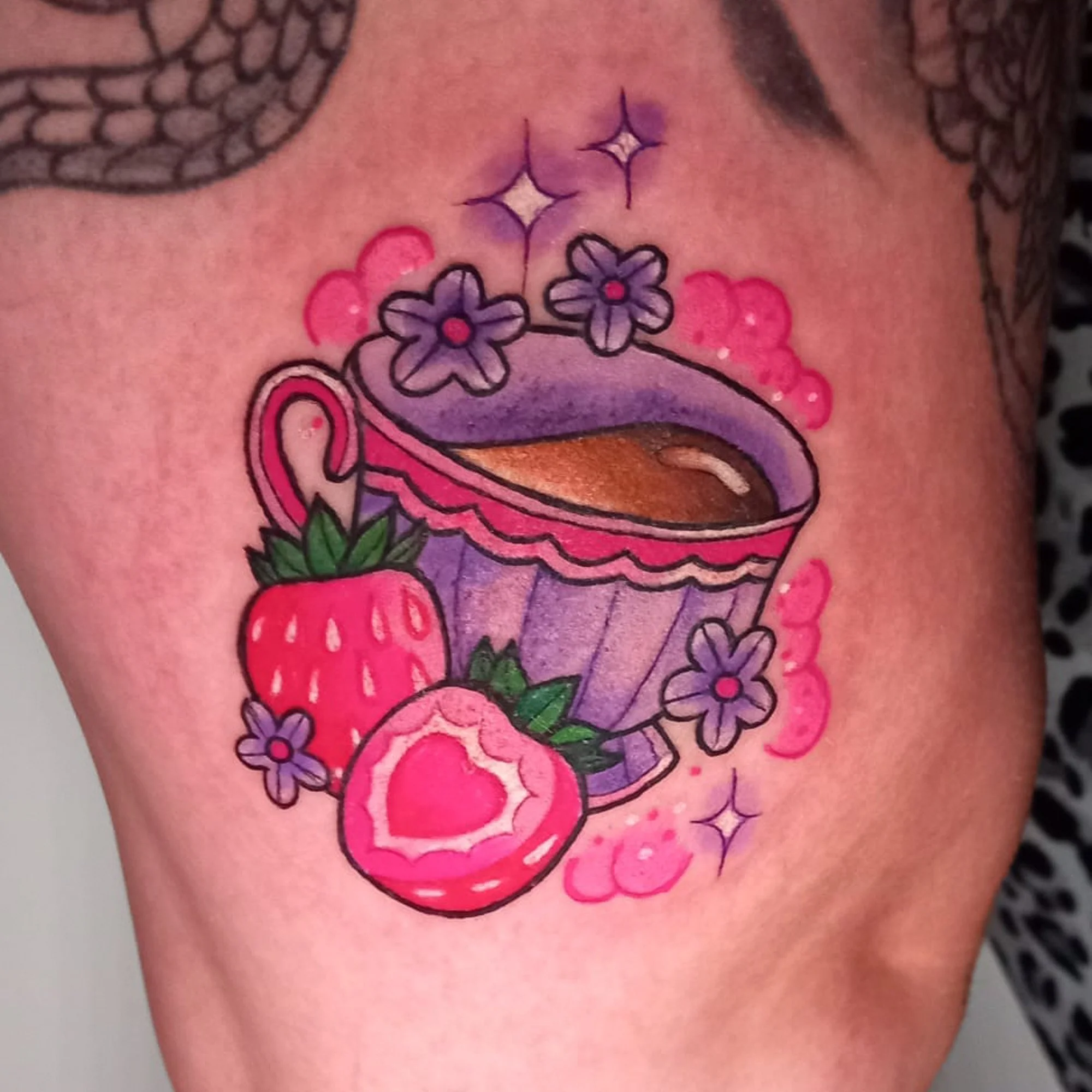 Colorful tattoo of a cupcake topped with purple frosting and surrounded by pink hearts and purple flowers.