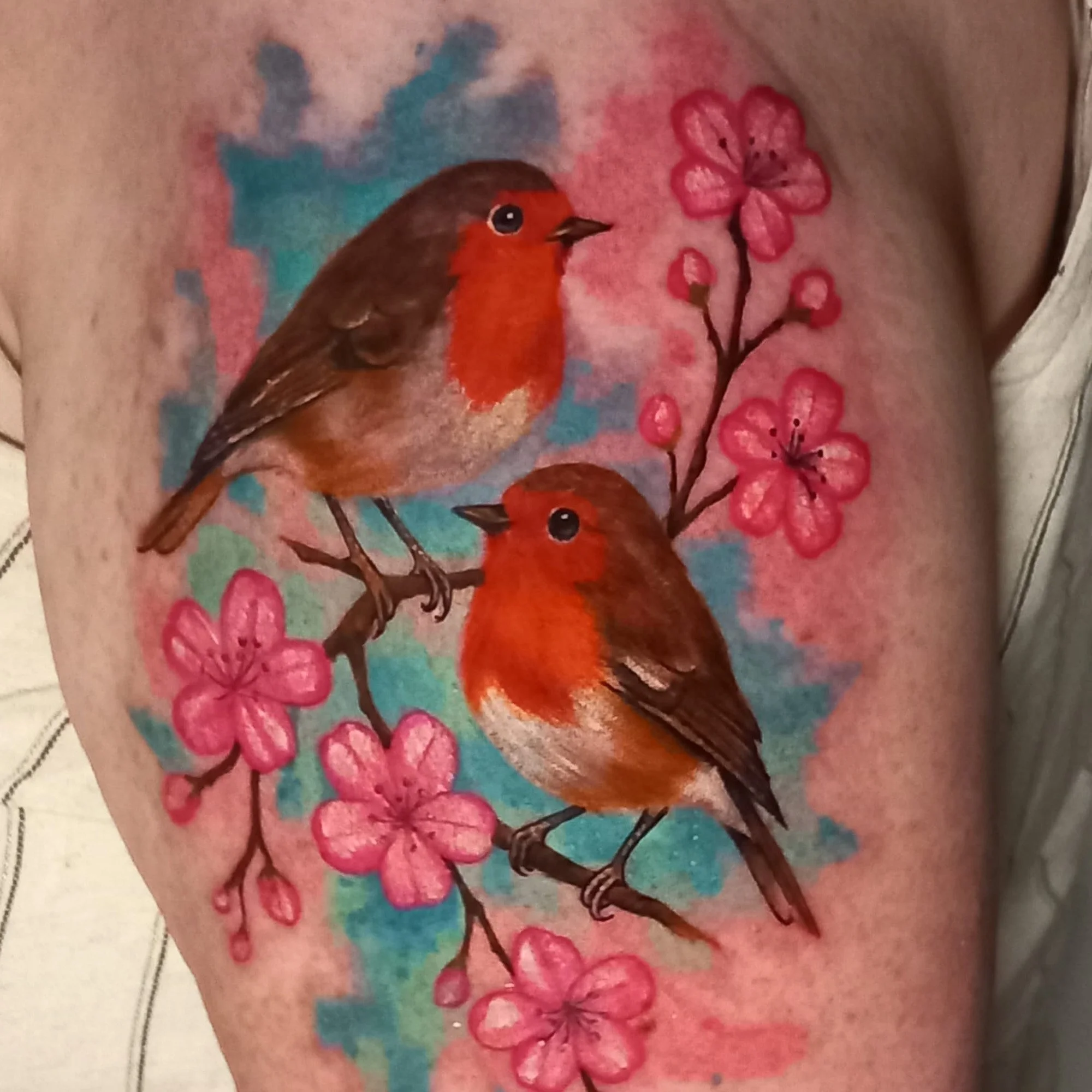 Tattoo of a bird on a person's side with pink and blue flowers and watercolor-style background.