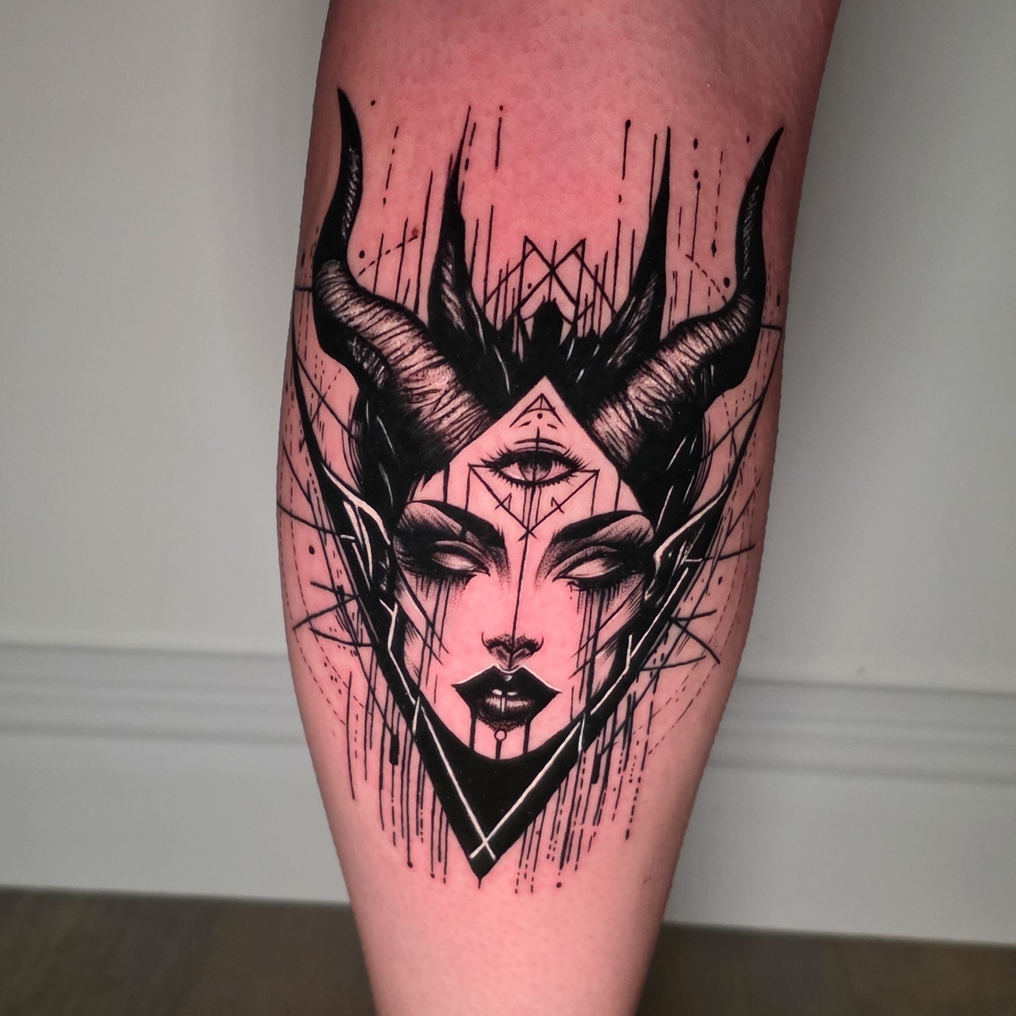 Tattoo of a woman's face with geometric design, horns, and an eye on the forehead, located on a person's arm.