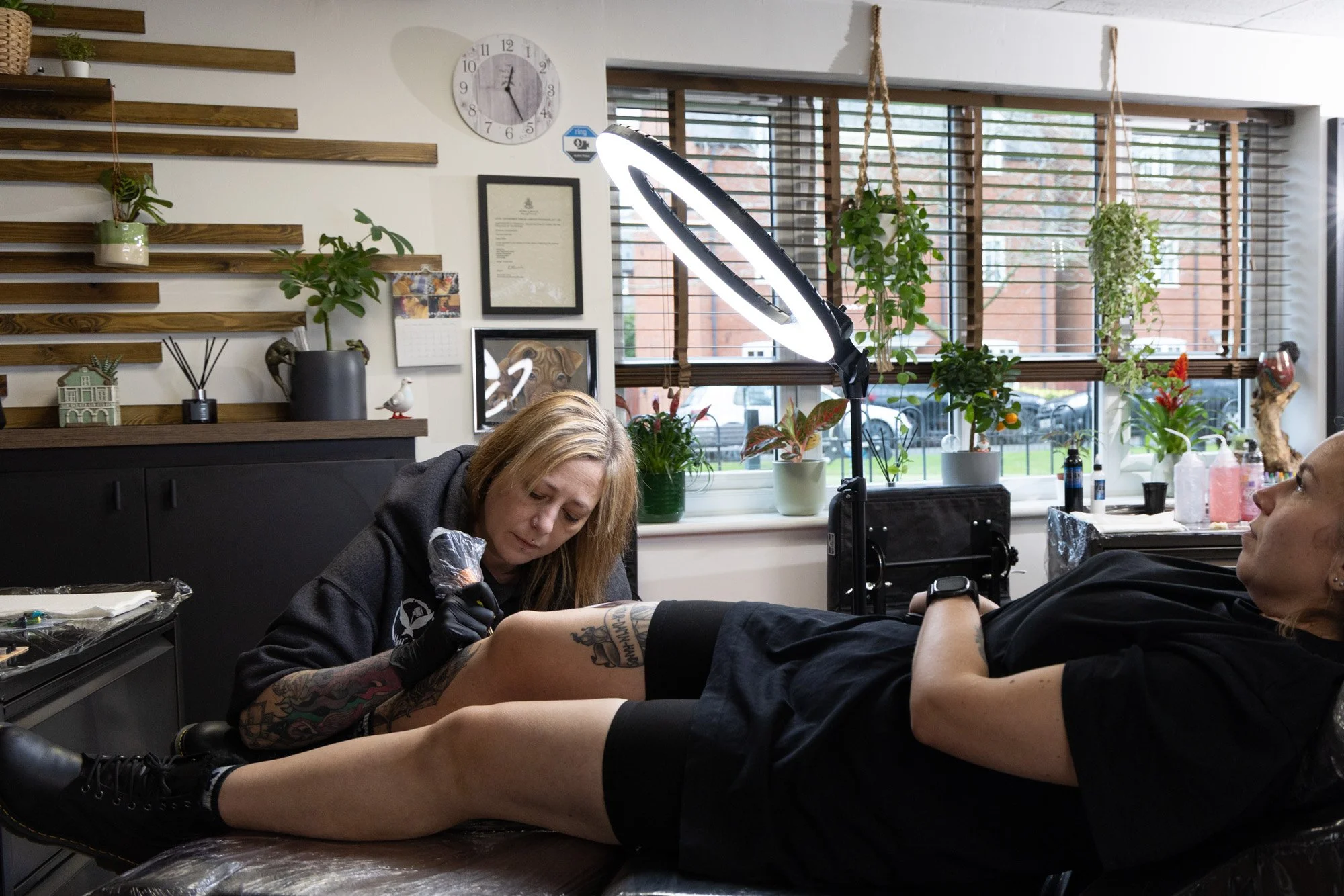A woman with tattoos getting a tattoo on her arm by an artist in a tattoo studio, with potted plants on display behind them and large windows in the background.