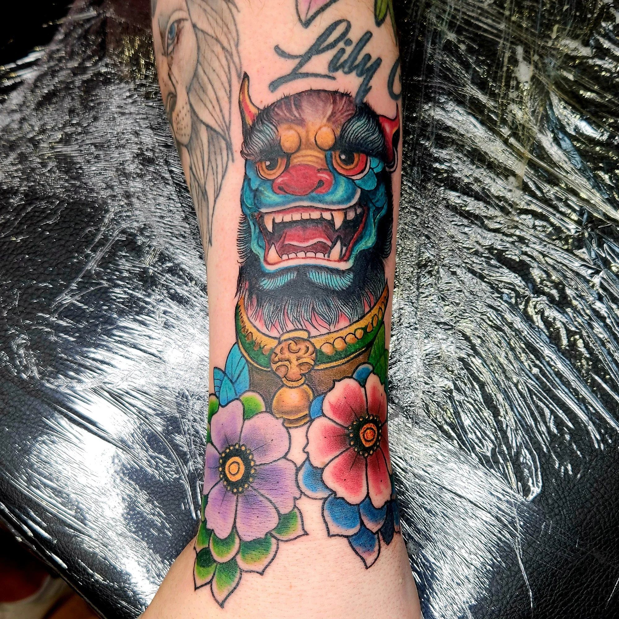 Colorful tattoo on forearm featuring a traditional Japanese-style demon mask with a red face, blue and green accents, and prominent fangs, alongside a handwritten word 'Lilly' at the top.