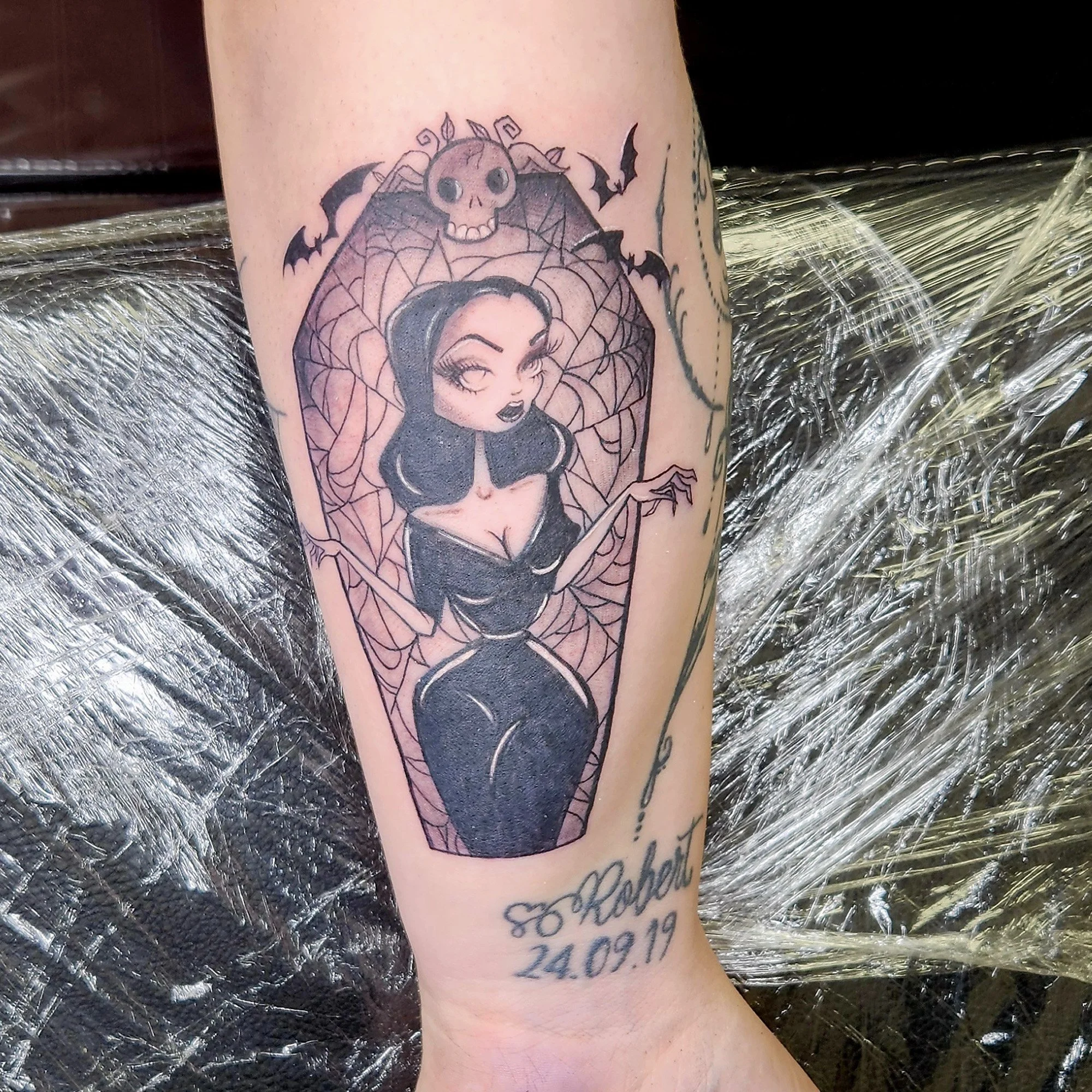 Tattoo of a cartoon woman with black hair in a spider web design, with small bats and a spider above her head, on a person's arm.