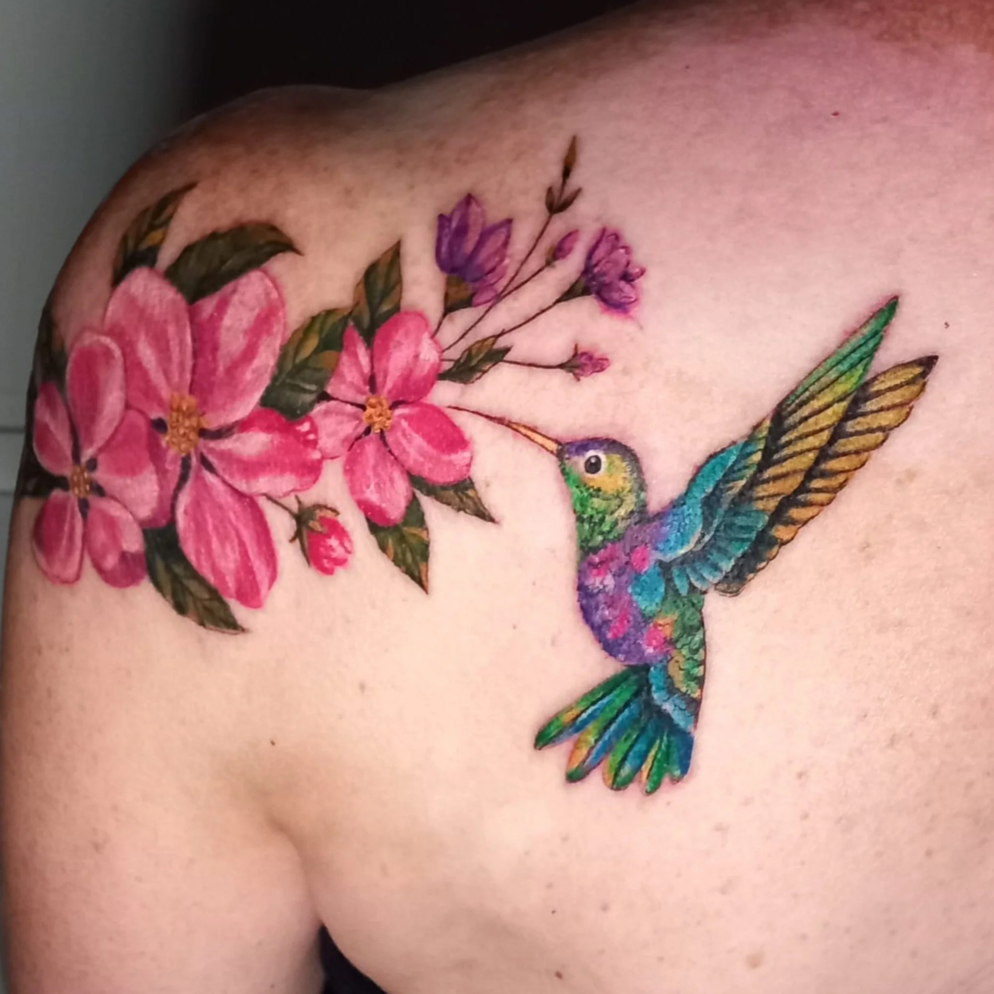 Colorful tattoo of vibrant pink flowers, purple flowers, and a bright blue and green hummingbird on skin.