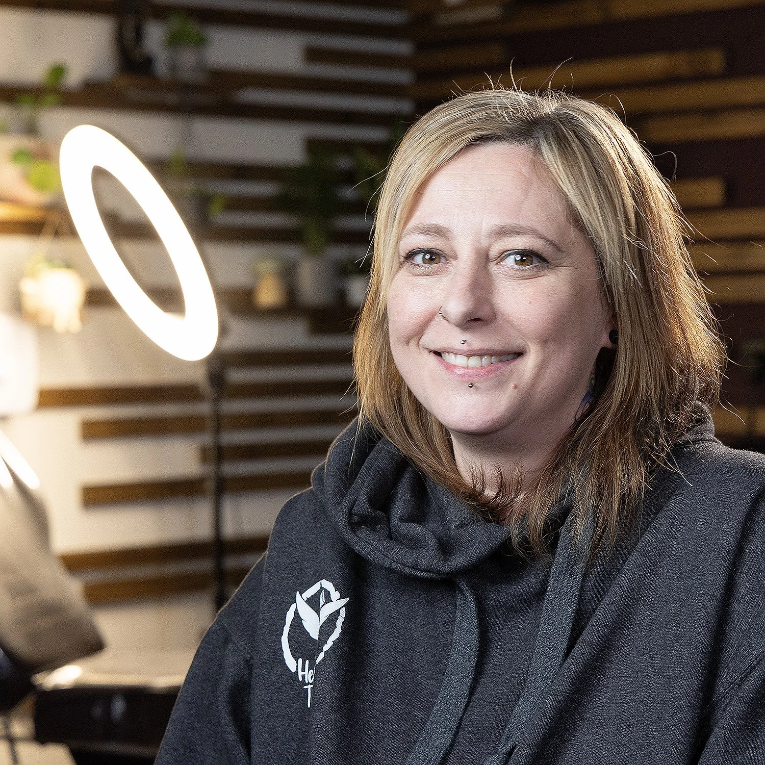A woman with blonde hair and piercings smiling. She is wearing a dark hoodie with a white logo on it. The background includes a modern wooden wall and a ring light.