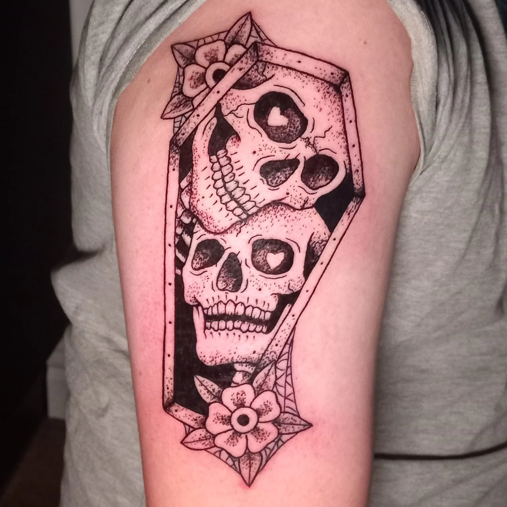 Tattoo of a skull with a flower and leaves inside a diamond-shaped frame on person's upper arm.