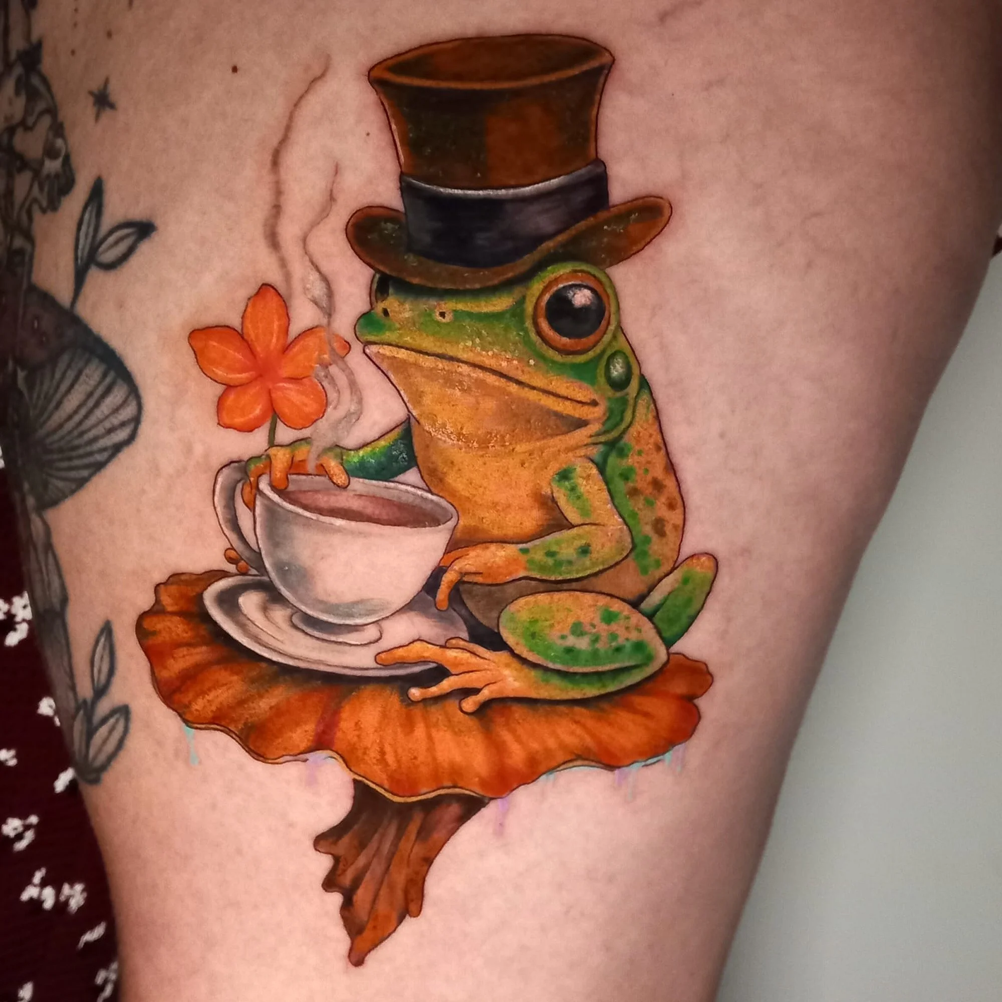 Tattoo of a colorful frog wearing a brown top hat, with an orange flower near it.