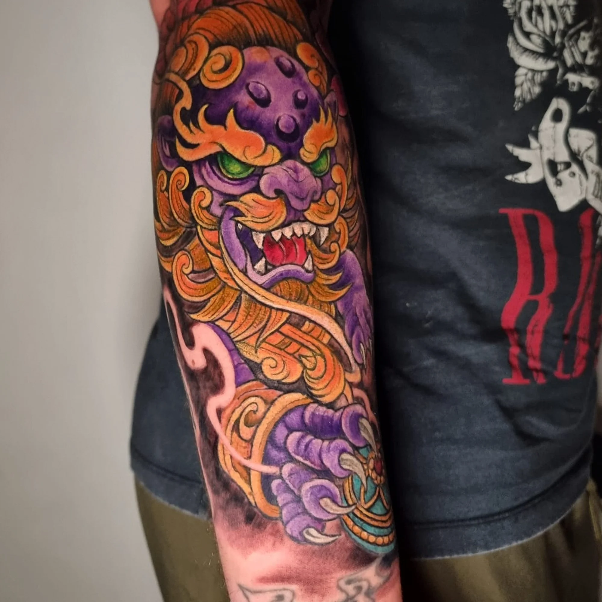 A vibrant, colorful tattoo of a fierce dragon with green eyes, orange and purple scales, and an open mouth showing sharp teeth, covering most of a person's arm.