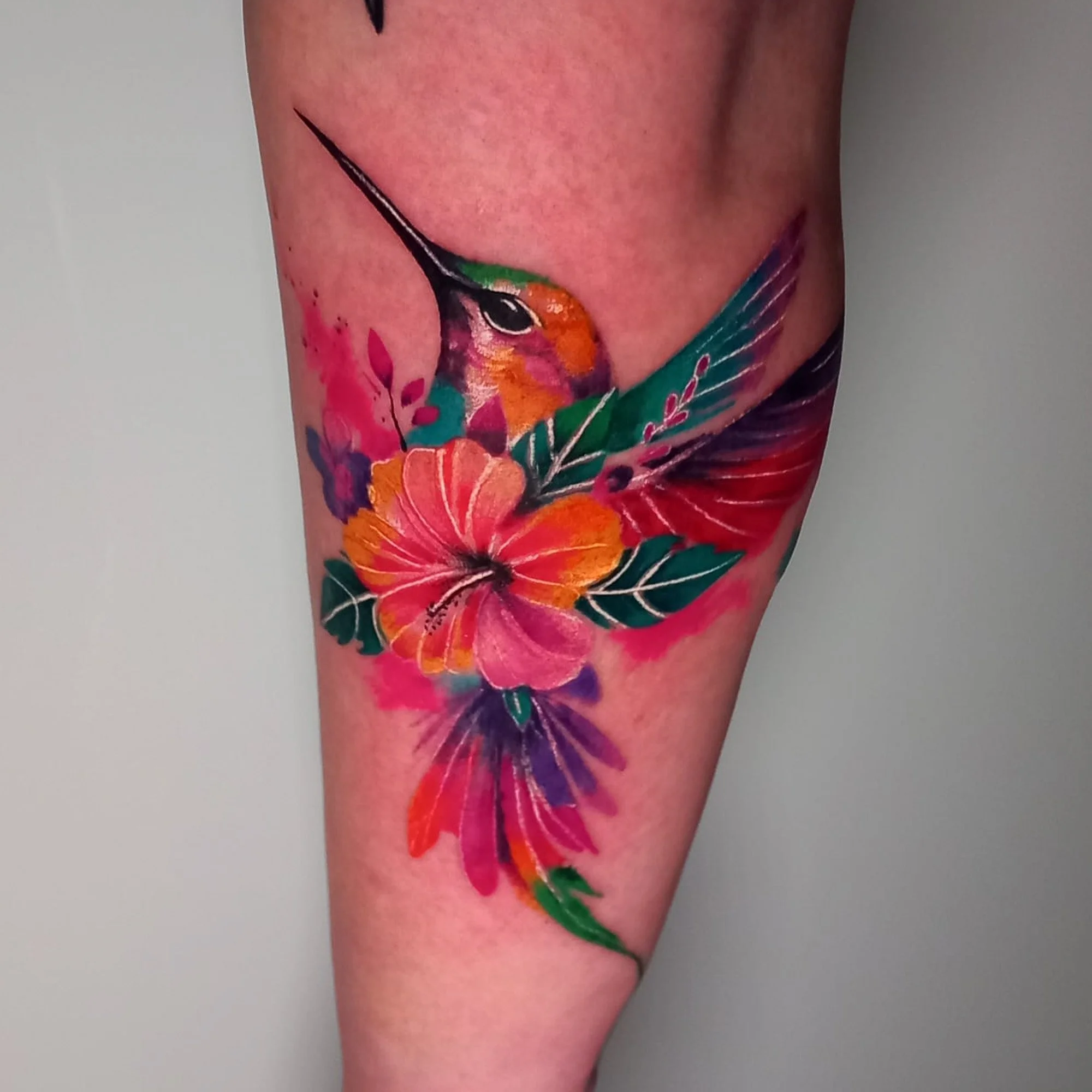 Close-up of a colorful hummingbird tattoo with vibrant pink, orange, purple, and green hues on a person's skin.