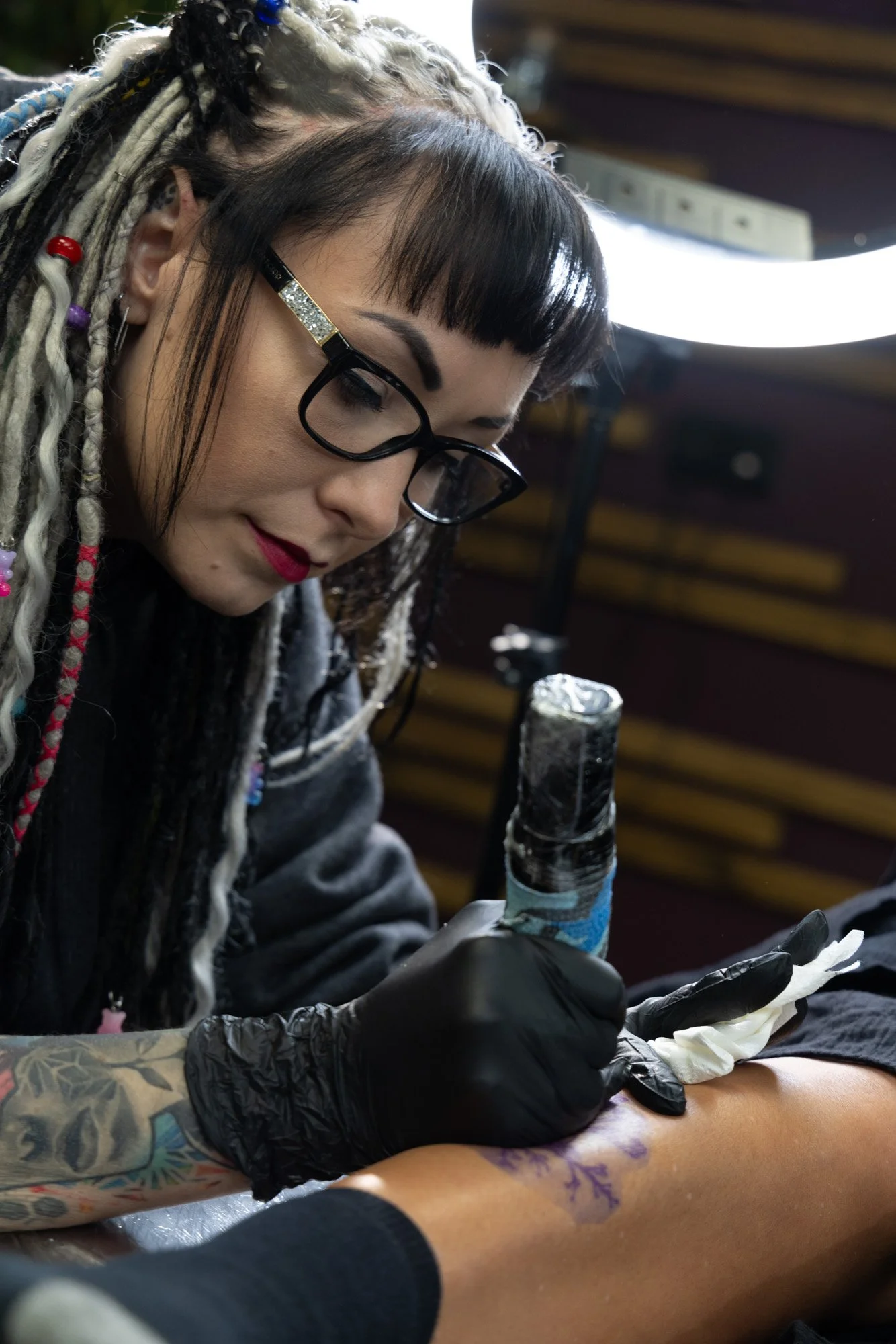 A woman with black glasses and black hair with bangs getting a tattoo on her forearm from an artist with multiple tattoos and dreadlocks. The artist is wearing black gloves and is using a tattoo machine.