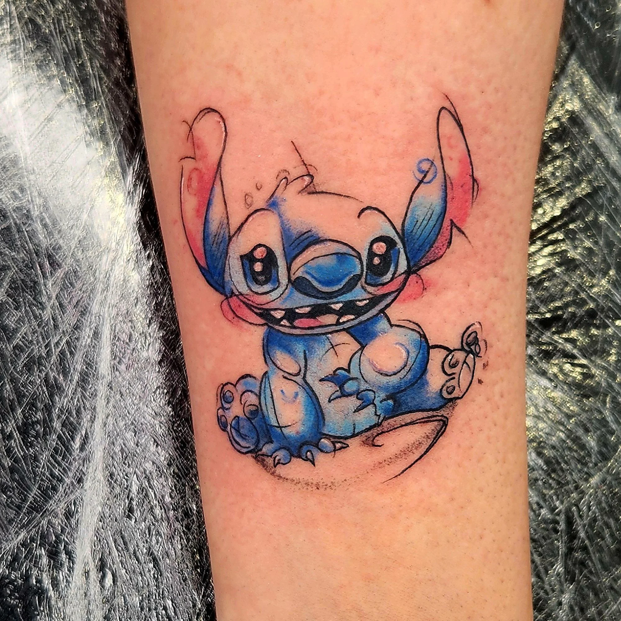 Colorful tattoo of a cartoon Stitch with big eyes on a person's arm.